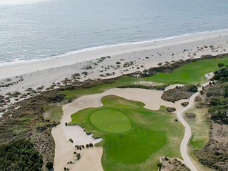An aerial view of a beach golf course with greens, fairways, sand traps, and surrounding dunes along the shoreline, under a hazy sky.