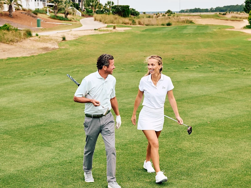 Two people walk golfing on a green course near houses, chatting and carrying clubs, sunny day, relaxed vibe.