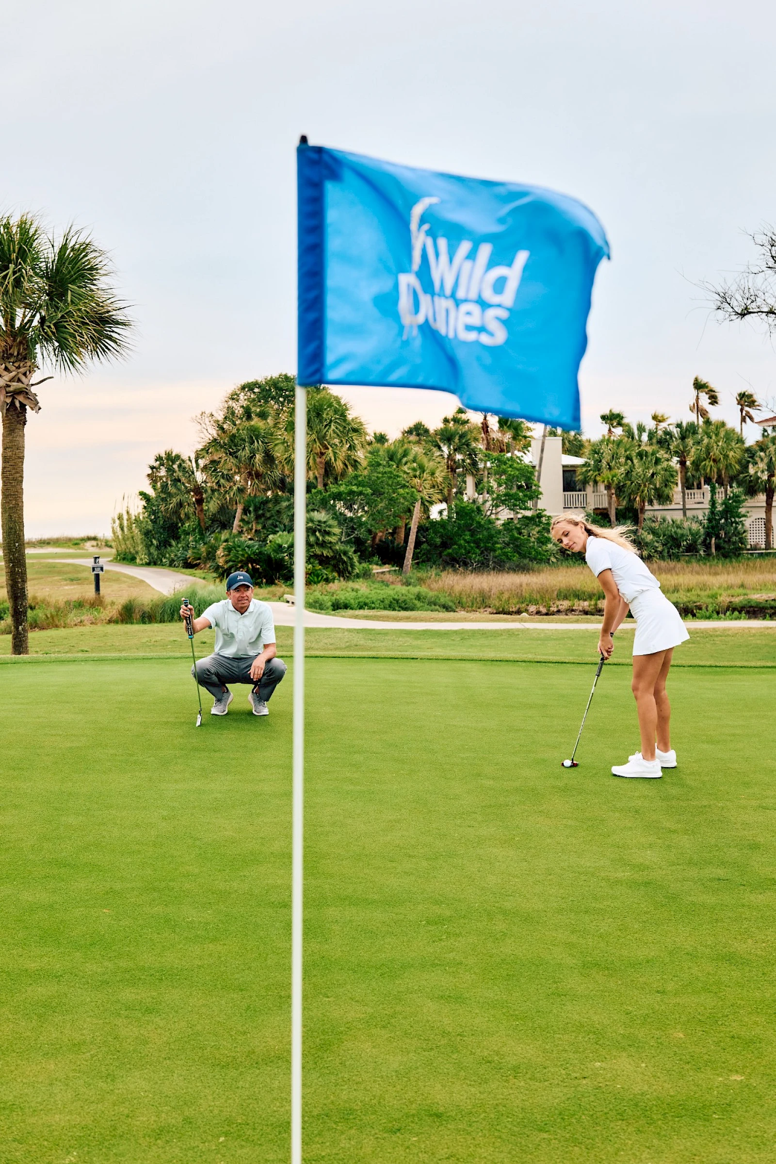 Two golfers on a sunny green, one putting near the hole while a teammate watches, with a blue Wild Dunes flag fluttering nearby.