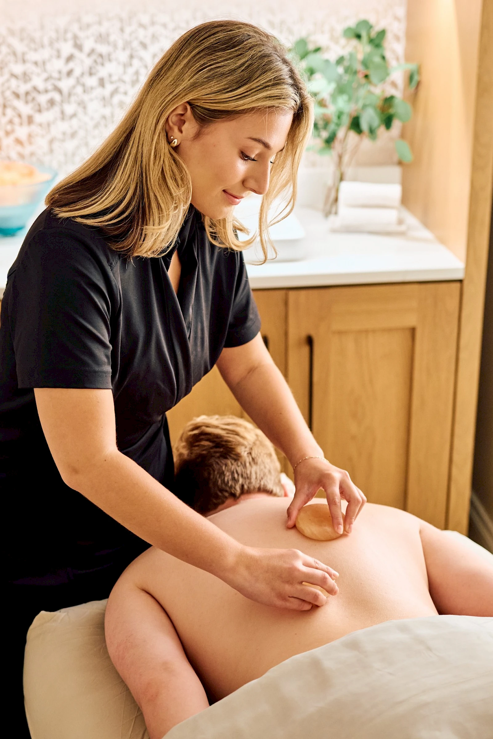A woman gives a back massage to a shirtless person lying face down on a massage table in a calm room.