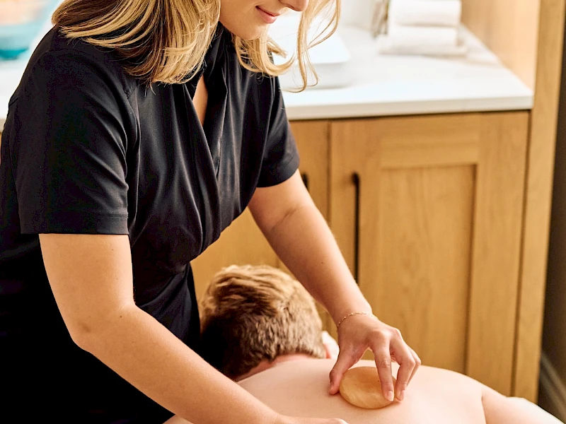 A woman gives a back massage to a shirtless person lying face down on a massage table in a calm room.
