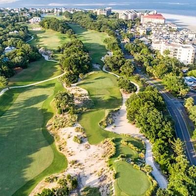 Aerial view of a coastal golf course with green fairways, sand traps, and palm trees, flanked by buildings and a blue ocean in the background.