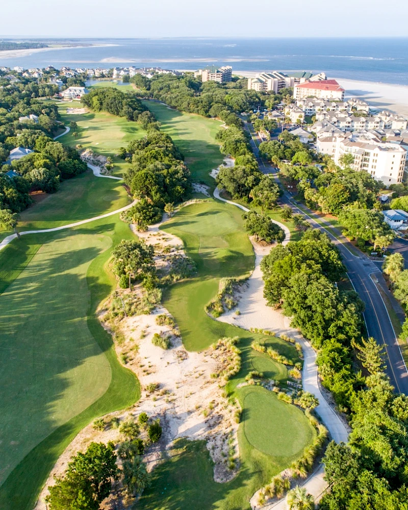 Aerial view of a coastal golf course with green fairways, sand traps, and palm trees, flanked by buildings and a blue ocean in the background.