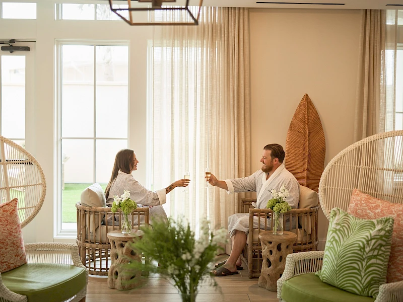 Two people sit in a bright, airy living room seaside-style, toasting with drinks while relaxing on woven chairs surrounded by plants and cushions.