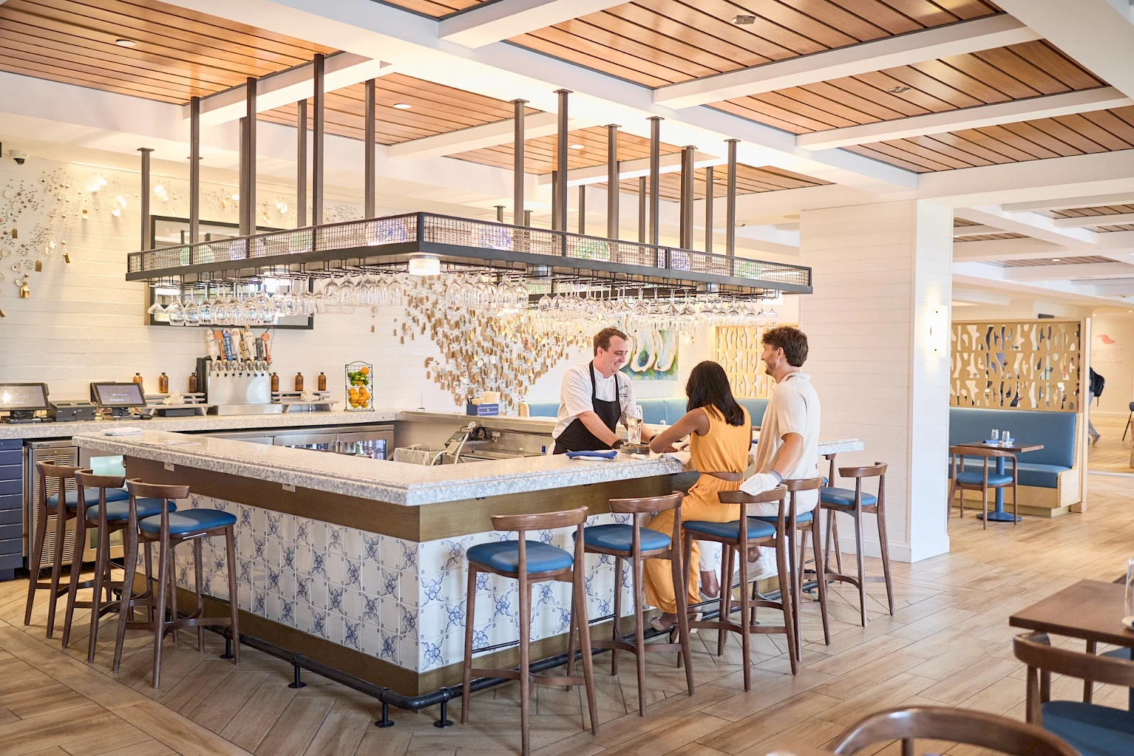 A stylish cafe or bar with a long counter, bar stools, and pendant lighting, where two staff serve two customers inside a modern dining area.