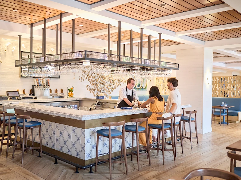 A stylish cafe or bar with a long counter, bar stools, and pendant lighting, where two staff serve two customers inside a modern dining area.