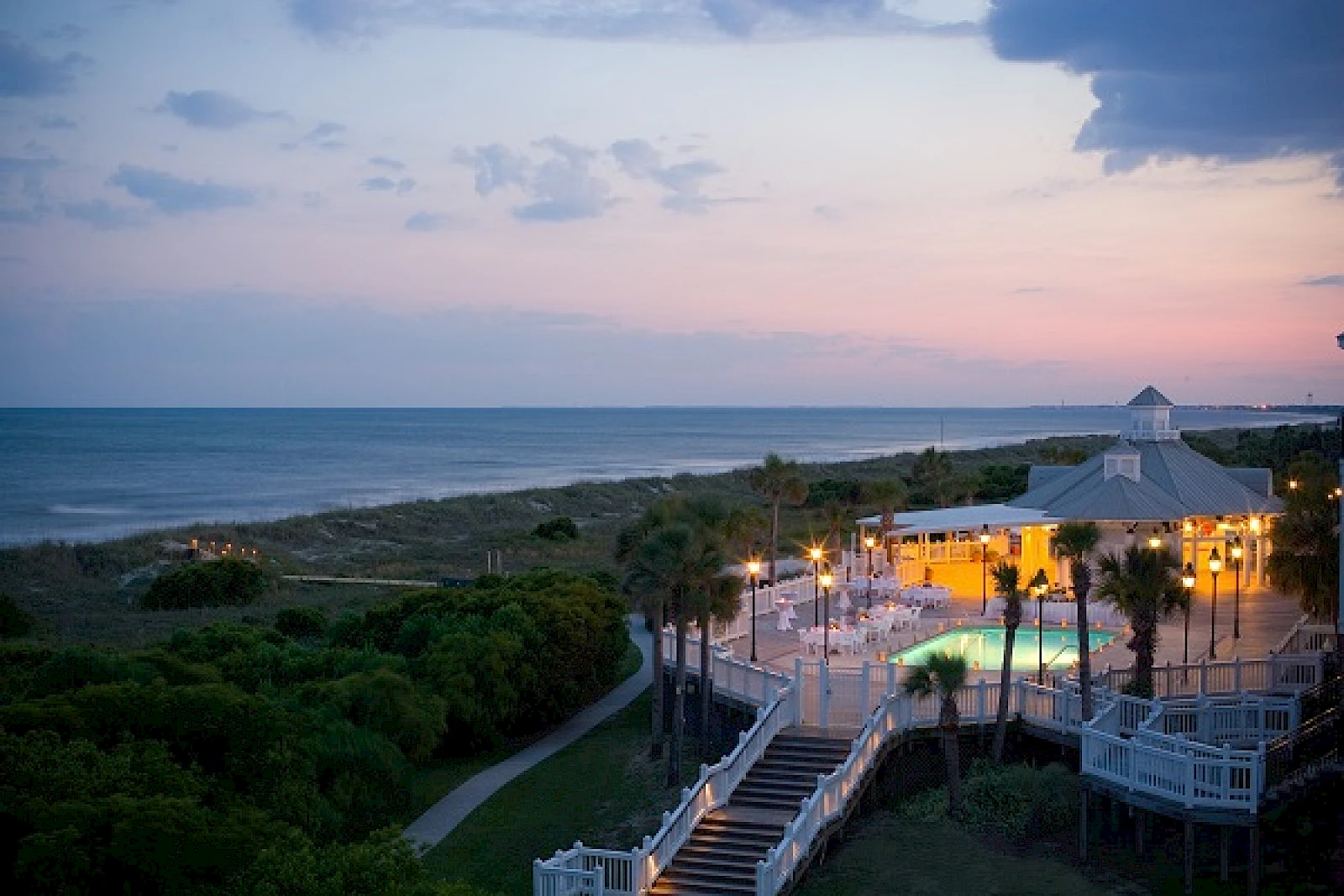 Beachfront scene at dusk with a boardwalk, palm trees, and a lit seaside venue overlooking the calm ocean, pastel sky, and gentle waves.