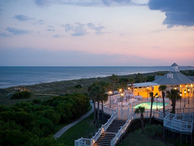 Beachfront scene at dusk with a boardwalk, palm trees, and a lit seaside venue overlooking the calm ocean, pastel sky, and gentle waves.
