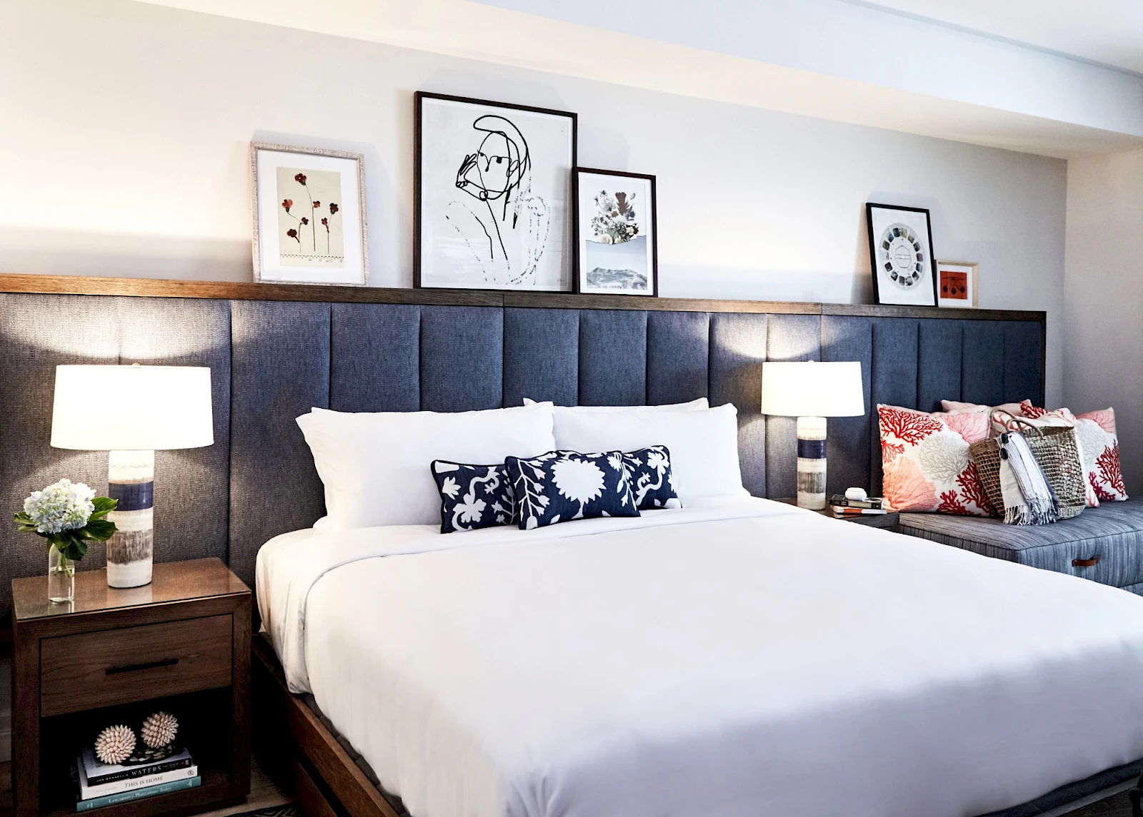 A modern hotel bedroom with a large white bed, dark upholstered headboard, two nightstands, matching lamps, framed art above the headboard, and a patterned throw pillow on a side chair, ending with a cozy vibe.