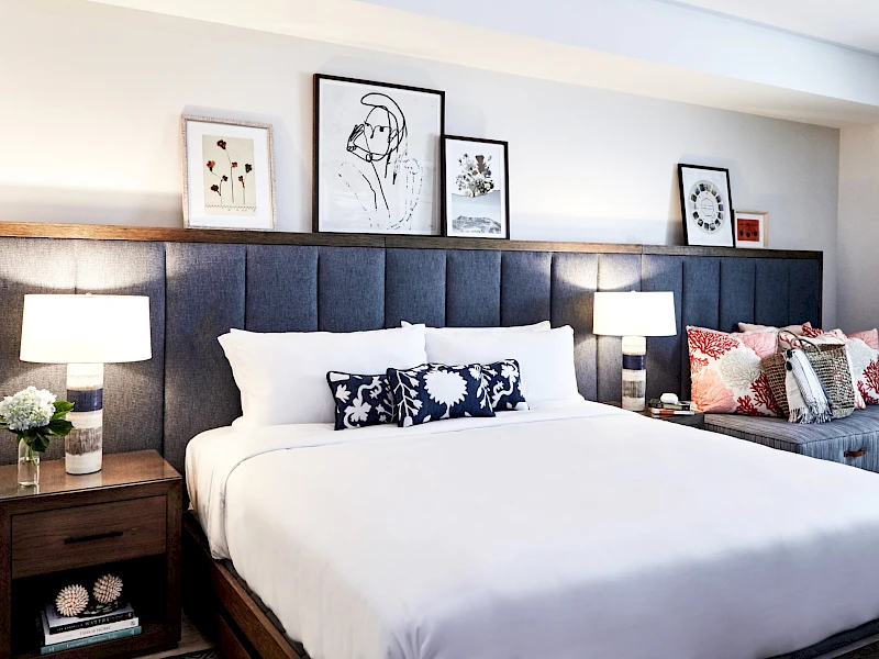 A modern hotel bedroom with a large white bed, dark upholstered headboard, two nightstands, matching lamps, framed art above the headboard, and a patterned throw pillow on a side chair, ending with a cozy vibe.