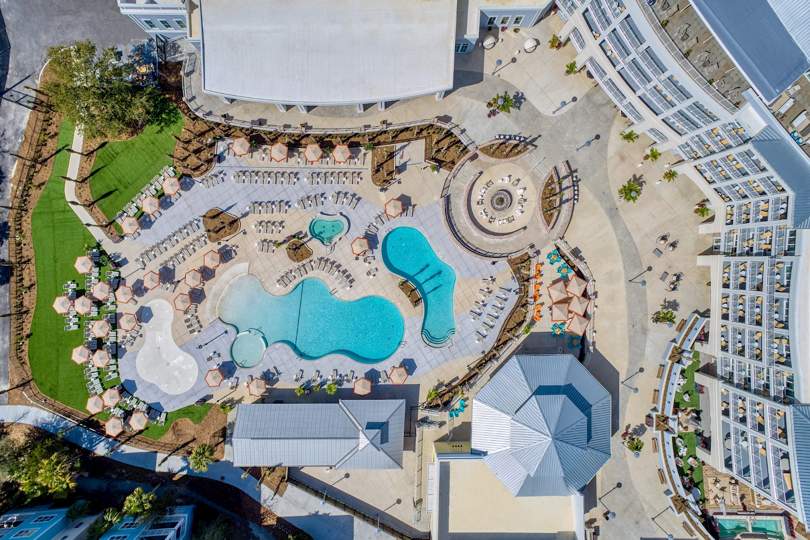 Aerial view of a resort pool area with a winding blue pool, lounge chairs, umbrellas, palm trees, surrounding buildings, and curved walkways, all sunny.