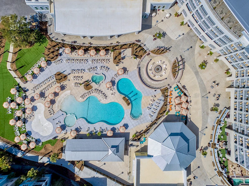 Aerial view of a resort pool area with a winding blue pool, lounge chairs, umbrellas, palm trees, surrounding buildings, and curved walkways, all sunny.