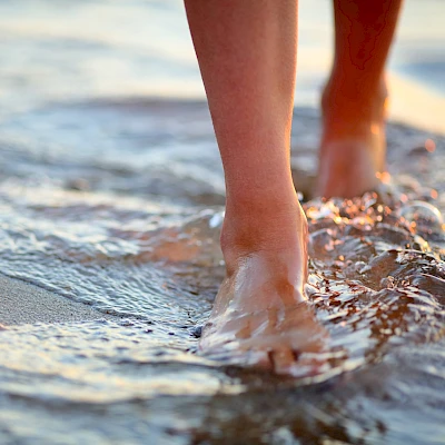 A person walking along a sandy beach with waves brushing their feet, footprints fading in foamy water.