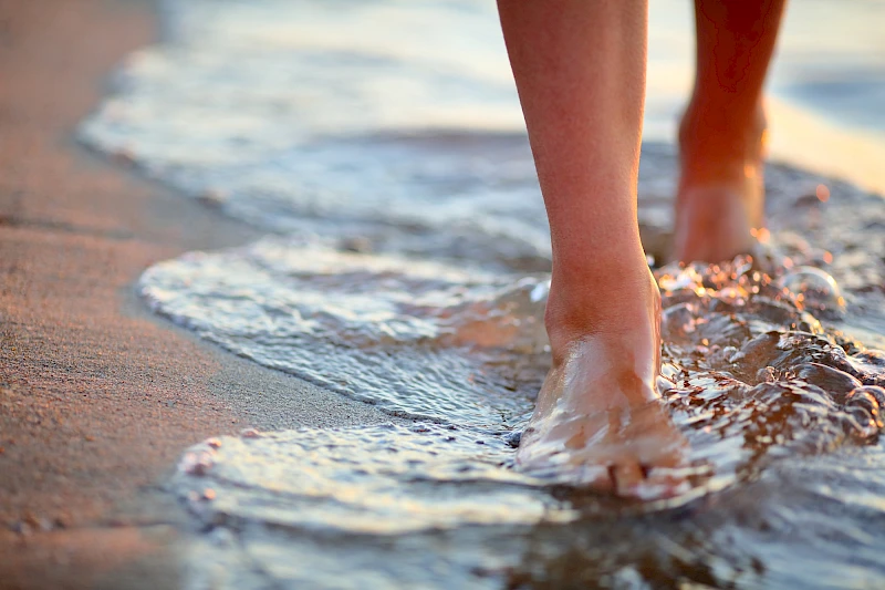 A person walking along a sandy beach with waves brushing their feet, footprints fading in foamy water.