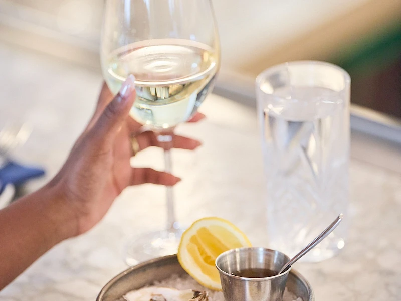 Someone holding a glass of white wine over a tray with lemon, oysters, and a metal cup with lemon wedge at a bar—cheers ending with a period.