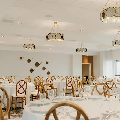 A bright banquet hall with round tables, white linens, and wooden chairs, set for a formal event with soft lighting.