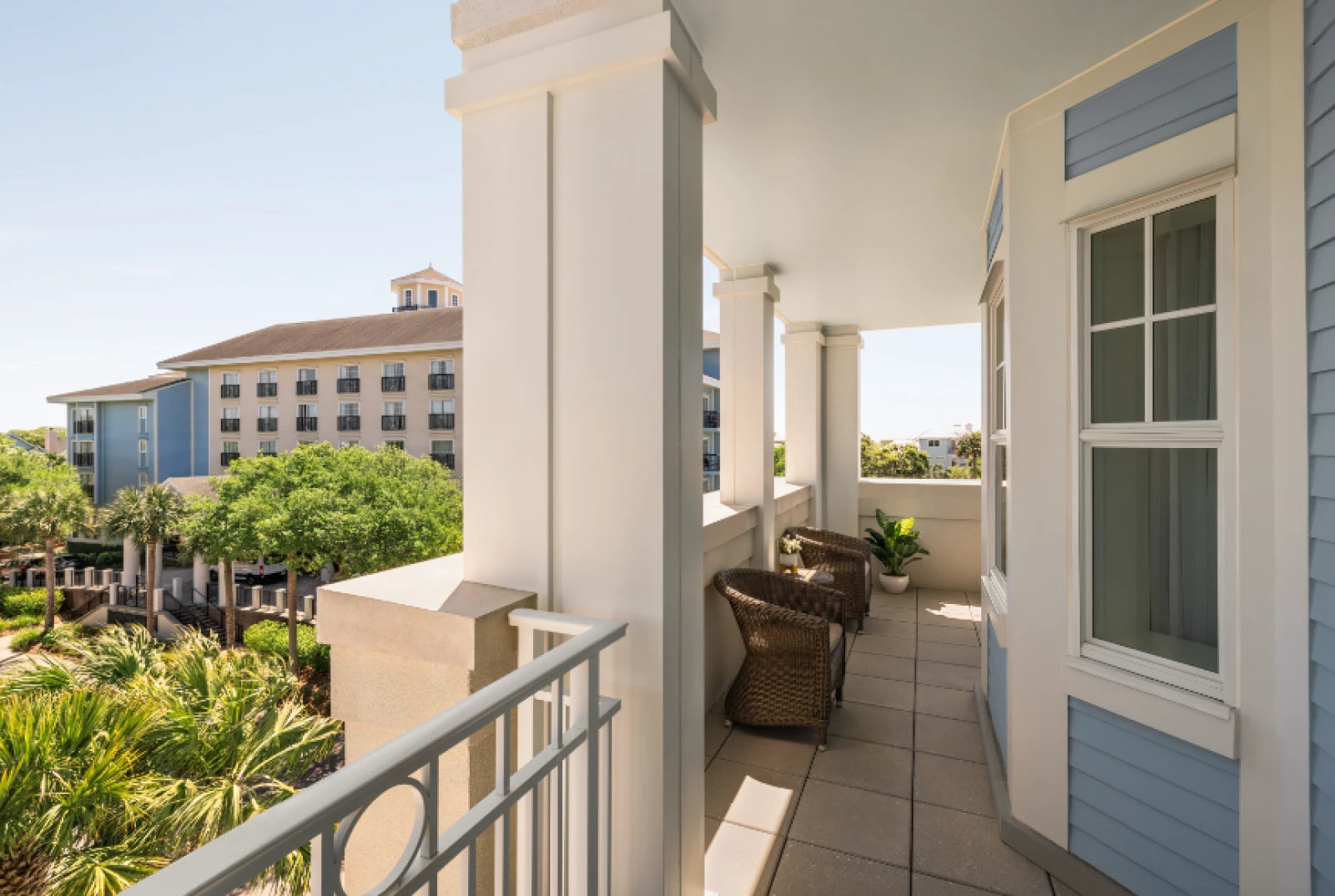 A bright, sunlit balcony with white columns and wicker chairs, overlooking a lush courtyard and neighboring buildings, peaceful coastal vibe.