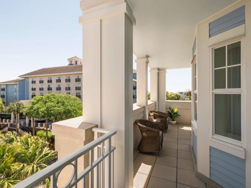 A bright, sunlit balcony with white columns and wicker chairs, overlooking a lush courtyard and neighboring buildings, peaceful coastal vibe.