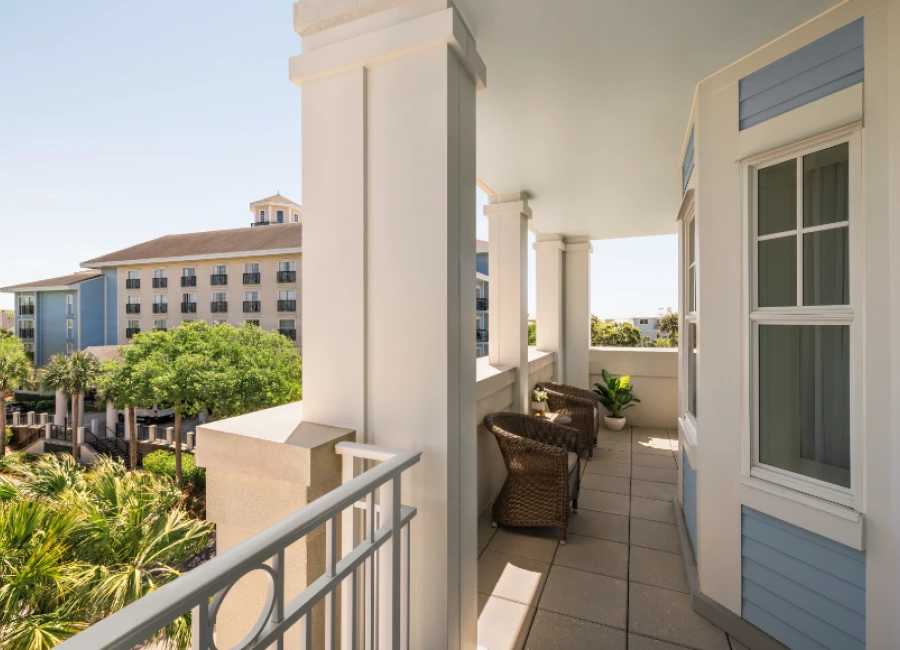 A bright, sunlit balcony with white columns and wicker chairs, overlooking a lush courtyard and neighboring buildings, peaceful coastal vibe.