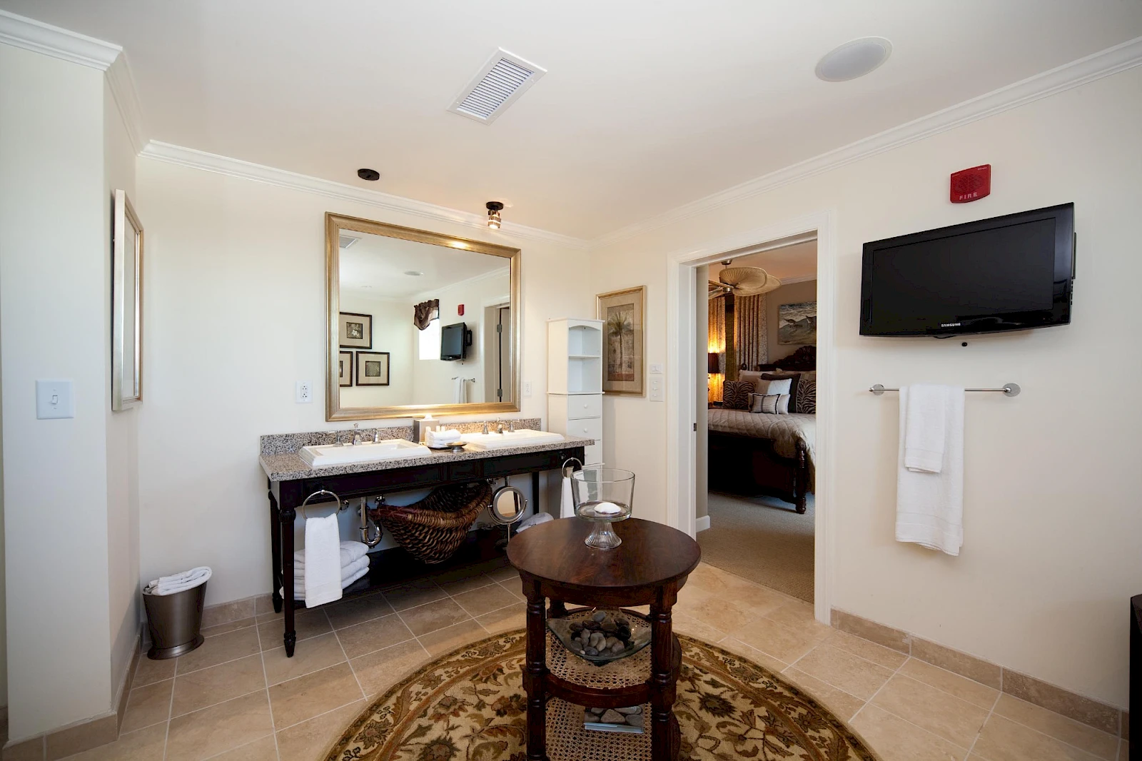 A bright hotel-style bathroom with a double sink vanity, large mirror, wall-mounted TV, towel rack, round rug, and a doorway to a bedroom; decor is clean and modern.