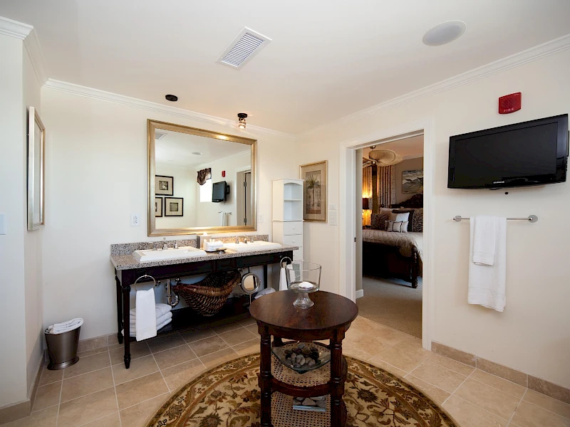 A bright hotel-style bathroom with a double sink vanity, large mirror, wall-mounted TV, towel rack, round rug, and a doorway to a bedroom; decor is clean and modern.