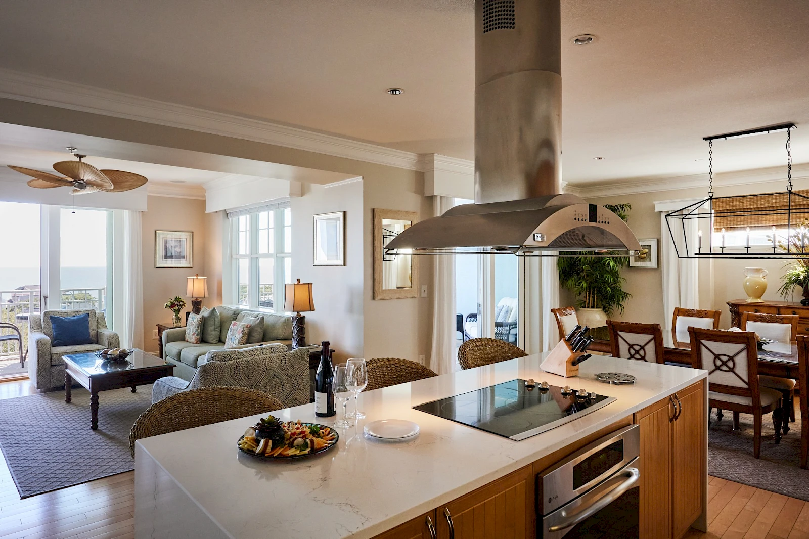 A modern open-plan kitchen and living area with an island stove, wicker seating, a dining area in the background, and a bright, airy vibe.