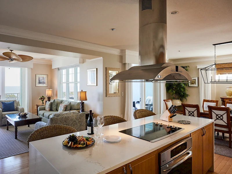 A modern open-plan kitchen and living area with an island stove, wicker seating, a dining area in the background, and a bright, airy vibe.