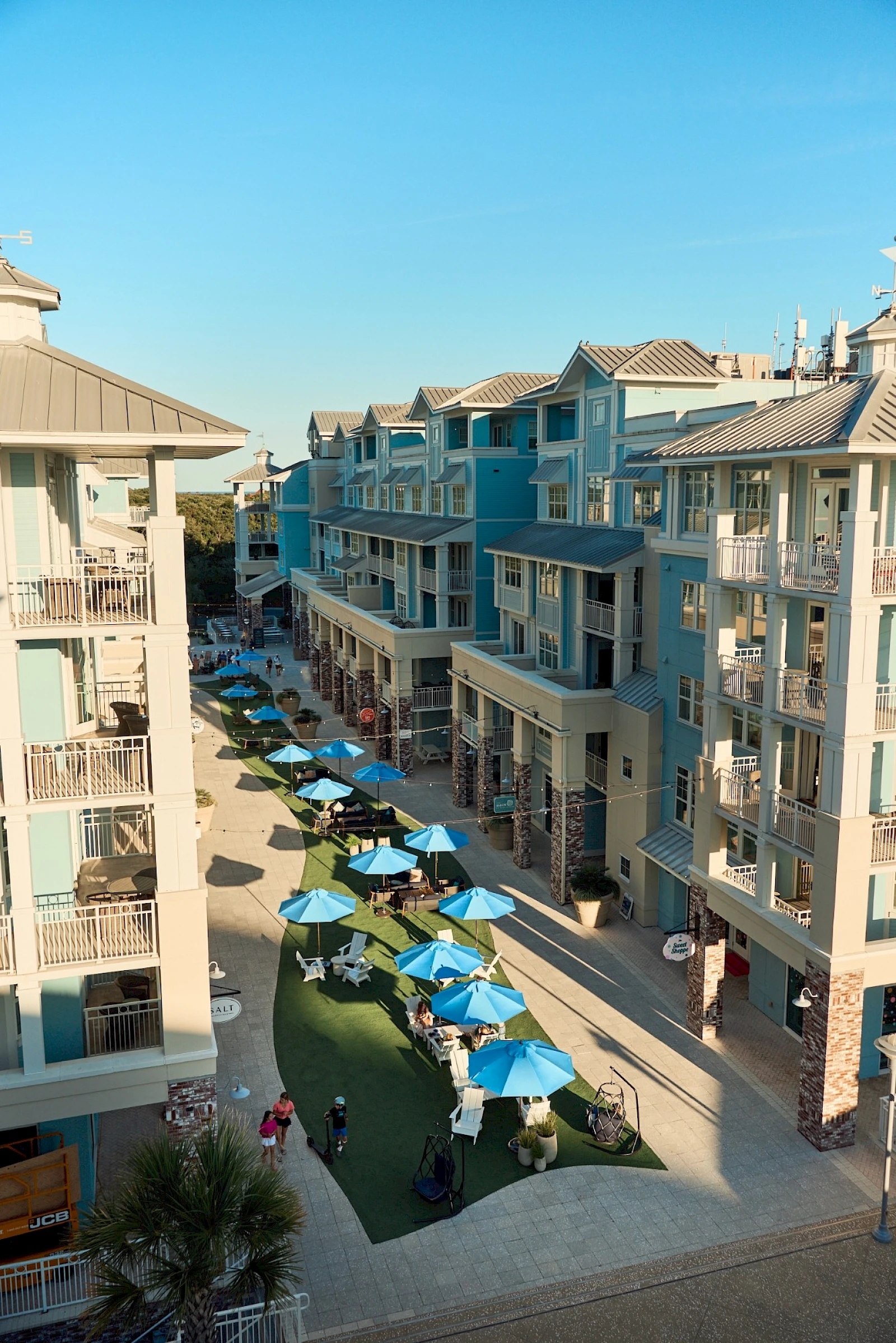 Aerial view of a sunny coastal mall lined with pastel blue buildings, outdoor seating under blue umbrellas, and people strolling along the pedestrian street.