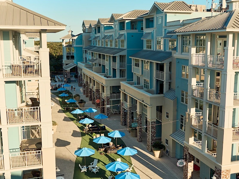 Aerial view of a sunny coastal mall lined with pastel blue buildings, outdoor seating under blue umbrellas, and people strolling along the pedestrian street.