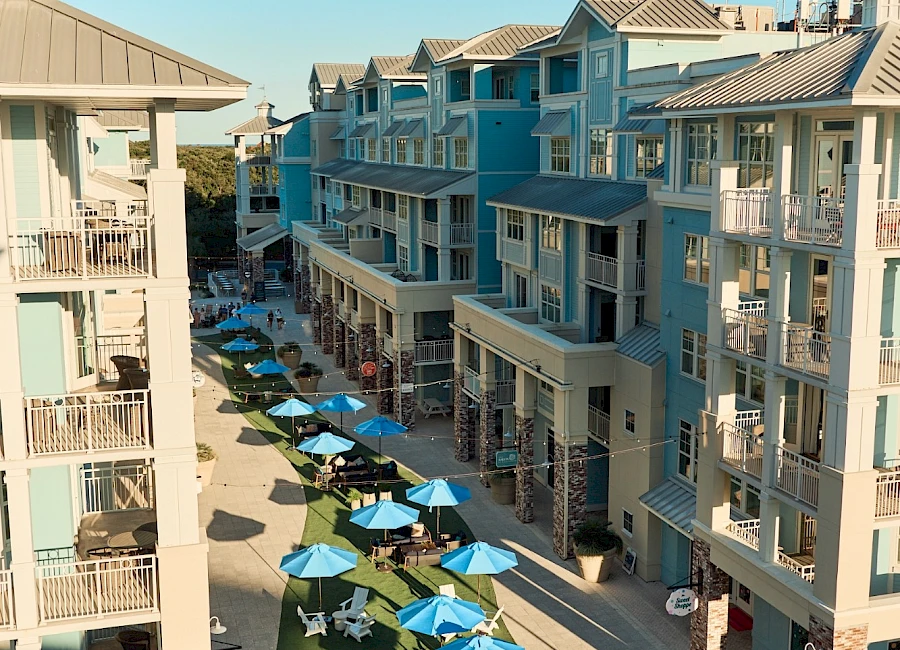 Aerial view of a sunny coastal mall lined with pastel blue buildings, outdoor seating under blue umbrellas, and people strolling along the pedestrian street.