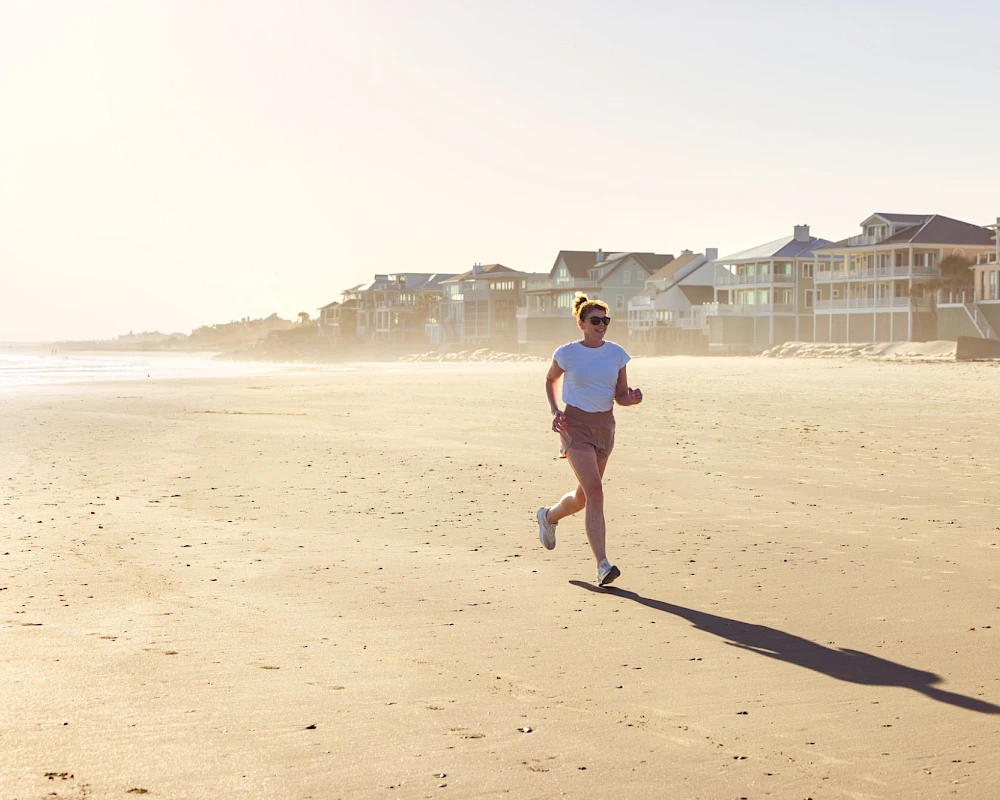 A man runs along a sunny beach with houses in the background and a long shadow trailing behind him. Running on the sand near the shoreline.