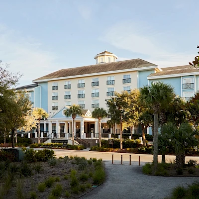 A light-blue hotel with a covered entrance sits among palm trees and a sandy landscape, under a clear sky.