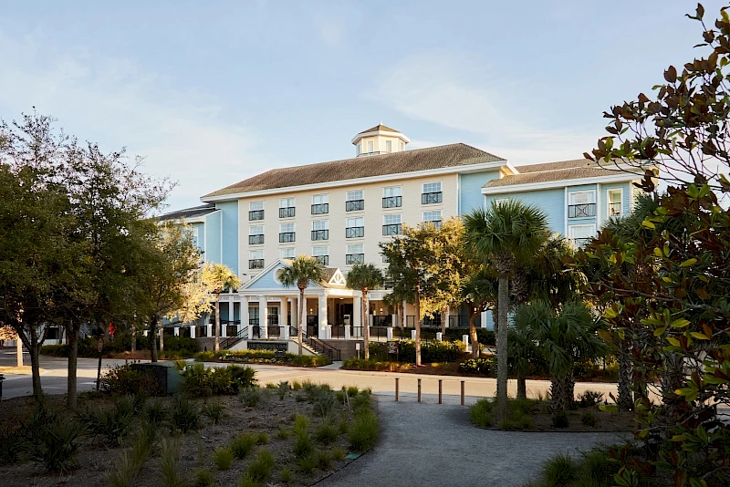 A light-blue hotel with a covered entrance sits among palm trees and a sandy landscape, under a clear sky.
