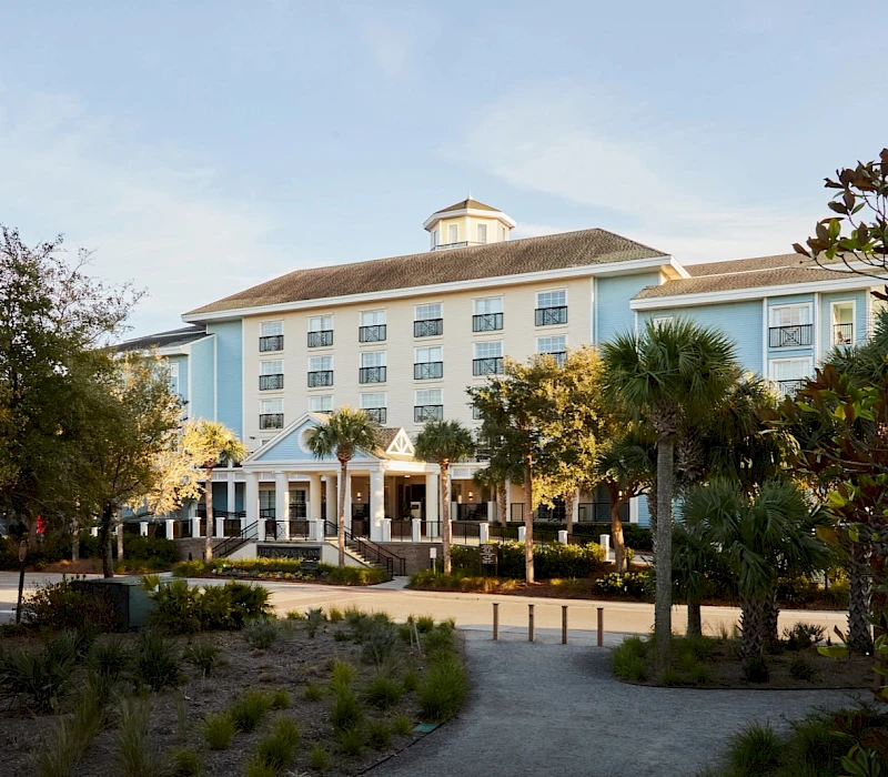 A light-blue hotel with a covered entrance sits among palm trees and a sandy landscape, under a clear sky.