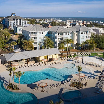 A resort-style pool area with palm trees, lounge chairs, and a large multi-story hotel complex under sunny skies by the coast.