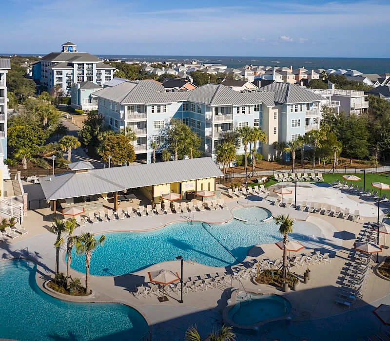 A resort-style pool area with palm trees, lounge chairs, and a large multi-story hotel complex under sunny skies by the coast.