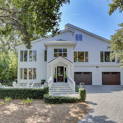 A large white two-story house with a gabled roof, multiple windows, a central front entrance, a brick walkway, and a two-car garage, set among trees.
