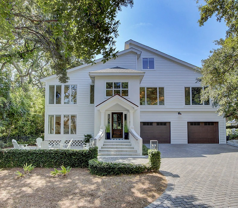 A large white two-story house with a gabled roof, multiple windows, a central front entrance, a brick walkway, and a two-car garage, set among trees.