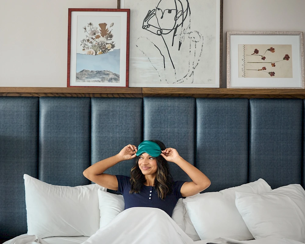 A woman sits up in bed with teal cap, smiling, under a white duvet; framed art lines the headboard wall above.