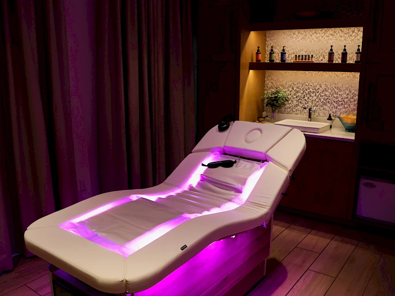 A spa treatment room with a reclining massage bed glowing purple, dim lighting, and a bar area in the background. The scene feels tranquil.
