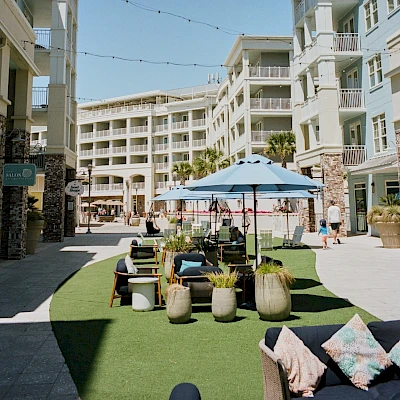 A sunny resort courtyard with multi-story white buildings, a central lawn, seating under umbrellas, and a few people walking around.