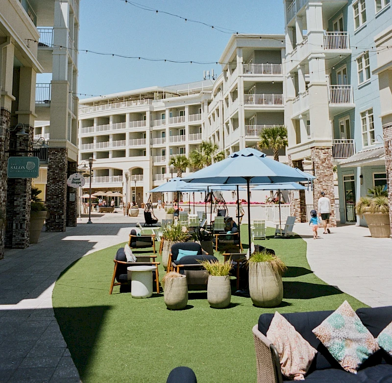 A sunny resort courtyard with multi-story white buildings, a central lawn, seating under umbrellas, and a few people walking around.