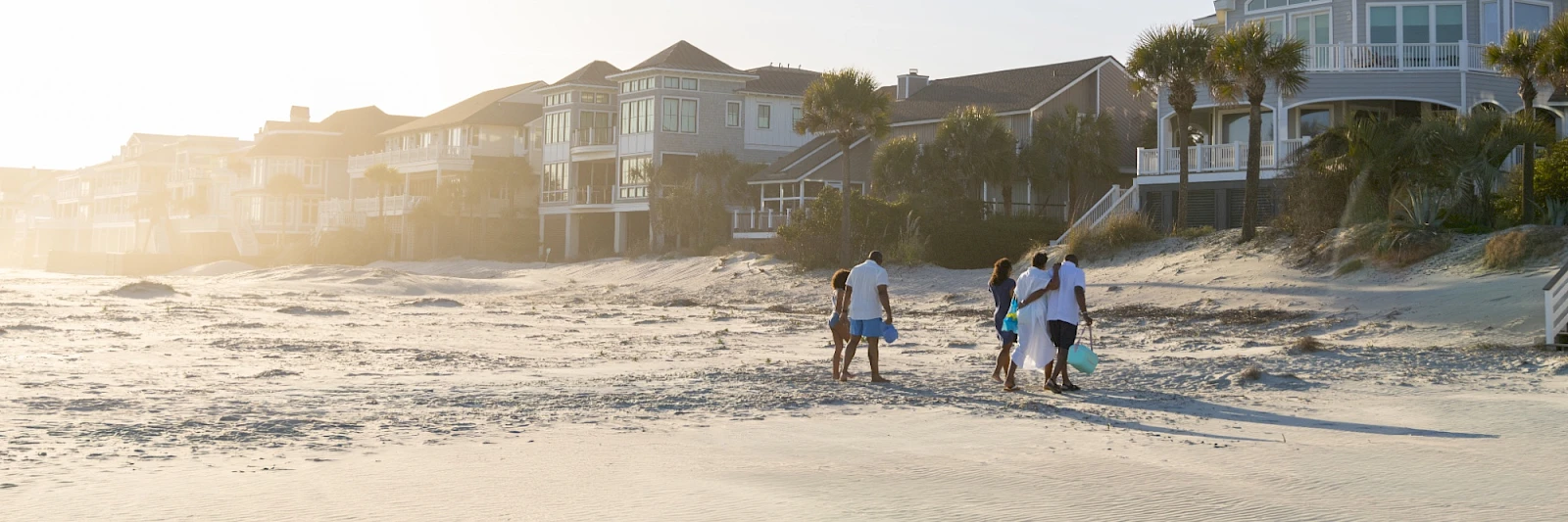 A family walks along a sunny beach near stylish houses, carrying a cooler and a bucket, as waves gently roll in.