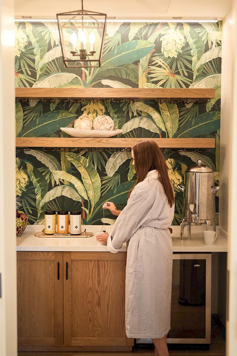 A person in a white robe stands at a kitchenette counter with jars and a drink dispenser, tropical leaf wallpaper behind, warm lighting.