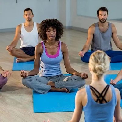 People in a yoga class sitting cross‑legged on mats, listening to an instructor at front.