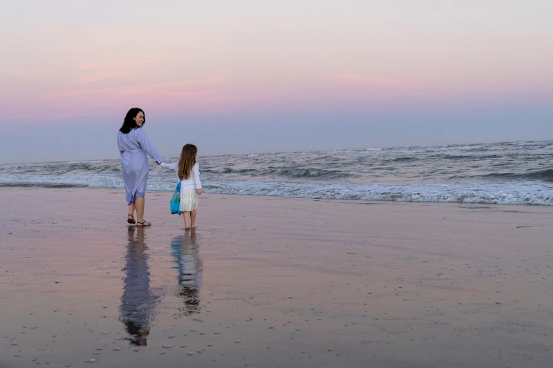 Two people walk along a calm beach at sunset, reflecting in the wet sand as gentle waves roll in, creating a peaceful, family moment.
