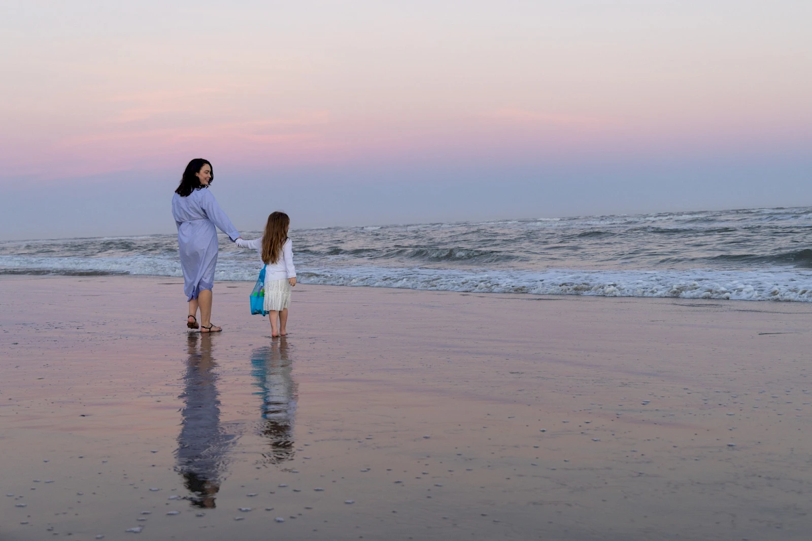 Two people walk along a calm beach at sunset, holding hands and reflected in the wet sand as gentle waves meet the shore.