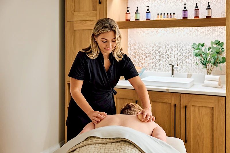 A spa scene with a massage therapist working on a client, a serene treatment room, warm wood cabinets, and neatly arranged bottles on a shelf.