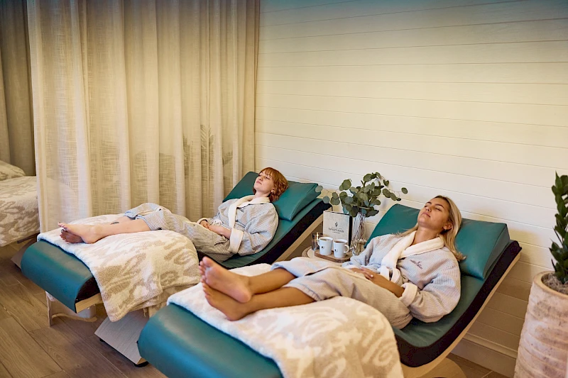 Two people are relaxing in recliner chairs at a spa, feet up under blankets, while a calm, cozy room with plants and soft lighting surrounds them.