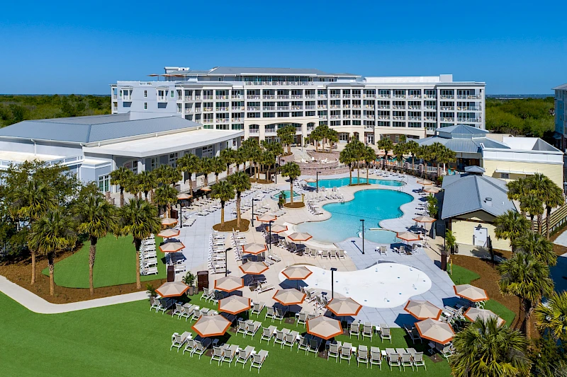 A large resort with a central pool area, sun loungers, palm trees, and multiple buildings in the background, all on a sunny waterfront setting.
